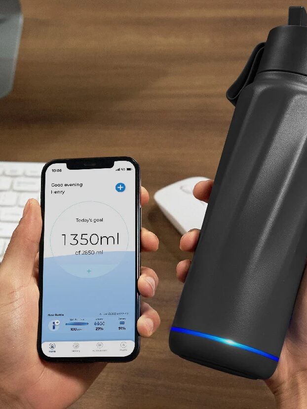 A person holds a smartphone displaying a hydration tracking app and a black smart waterbottle with a blue LED light, placed near a computer keyboard.