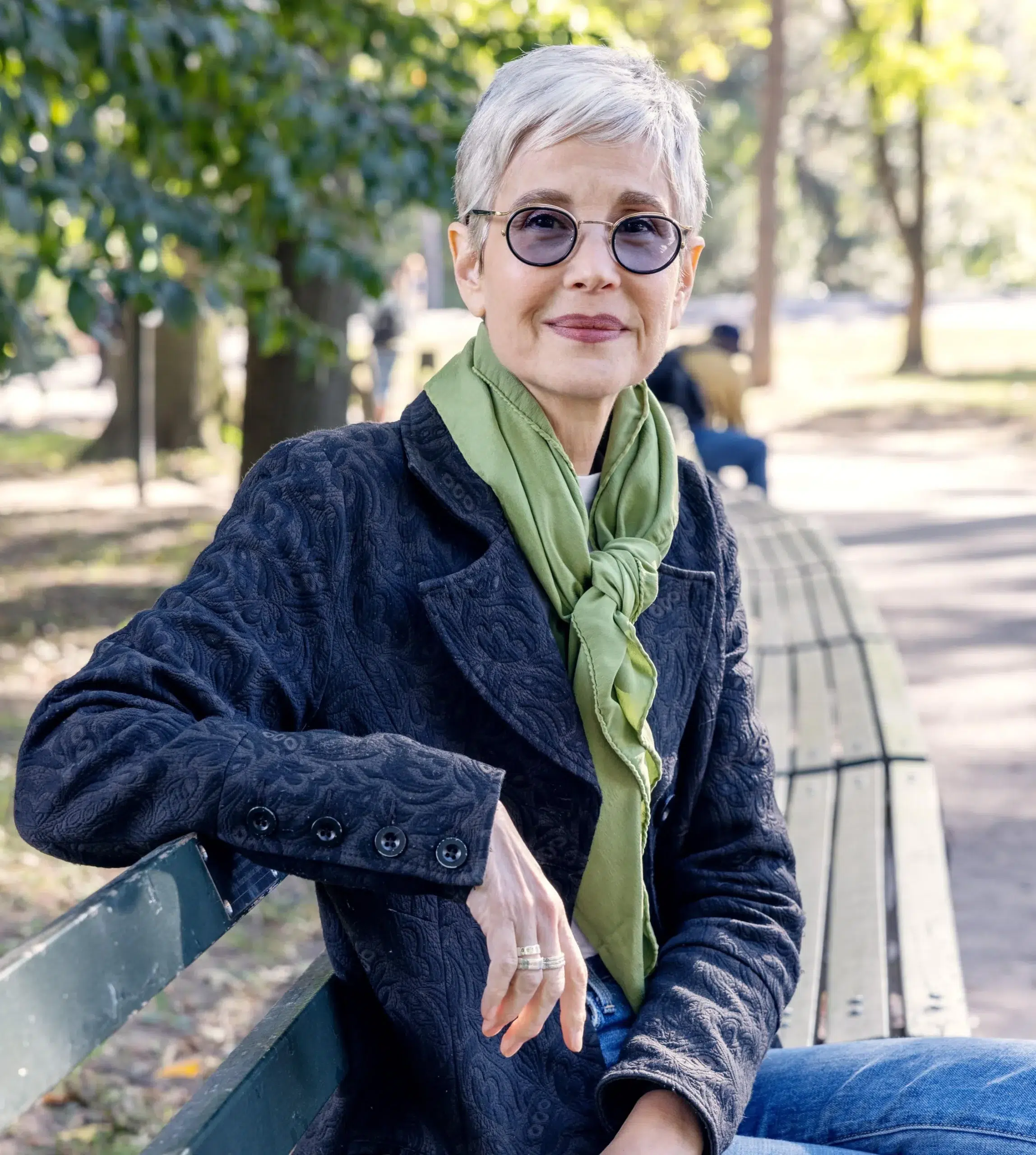 An older woman with short gray hair, glasses, and a green scarf sits on a park bench, looking at the camera. Trees and people are visible in the background.