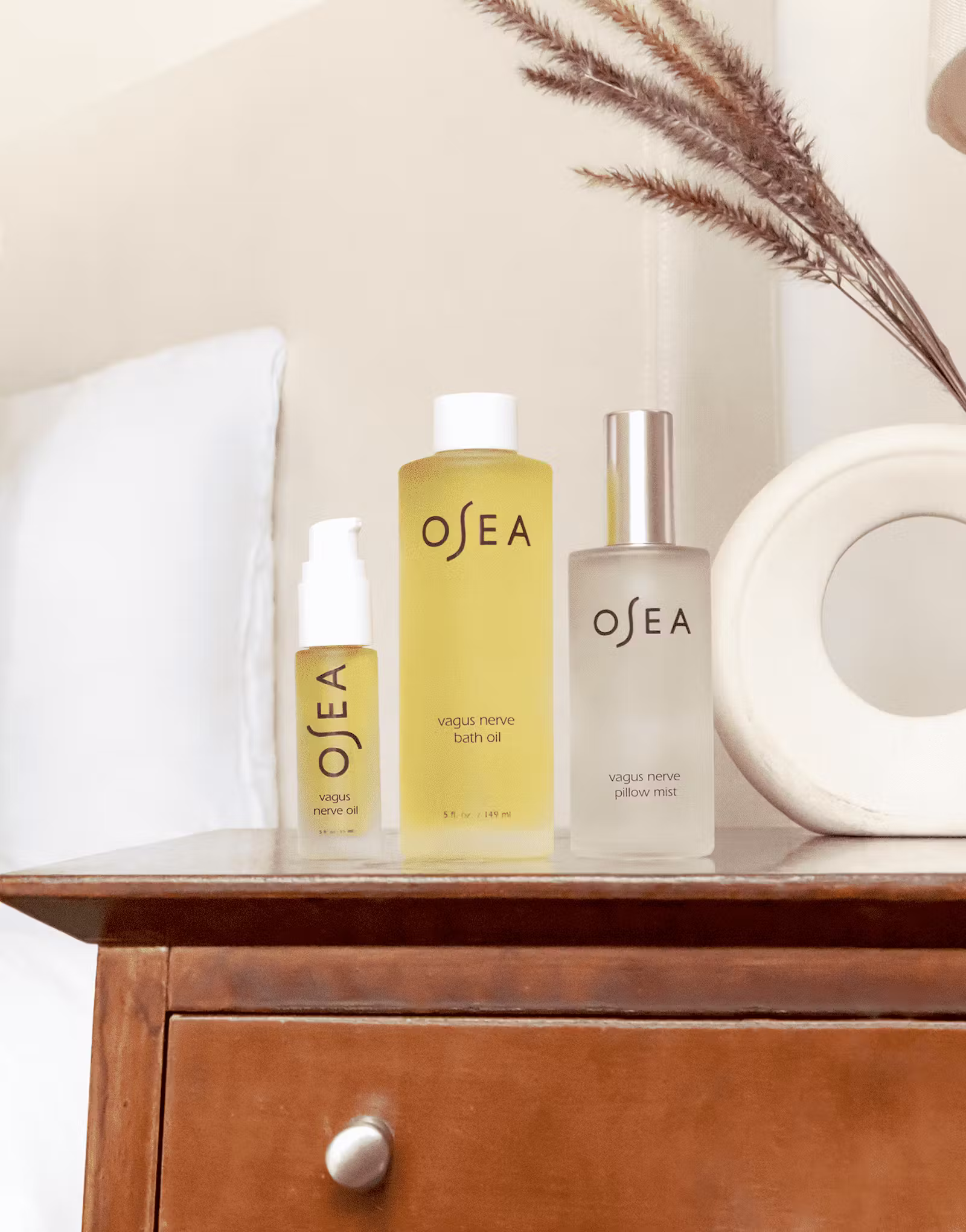 Three OSEA skincare products are arranged on a wooden nightstand next to a bed, including bath oil, pillow mist, and a serum, with decorative elements in the background.