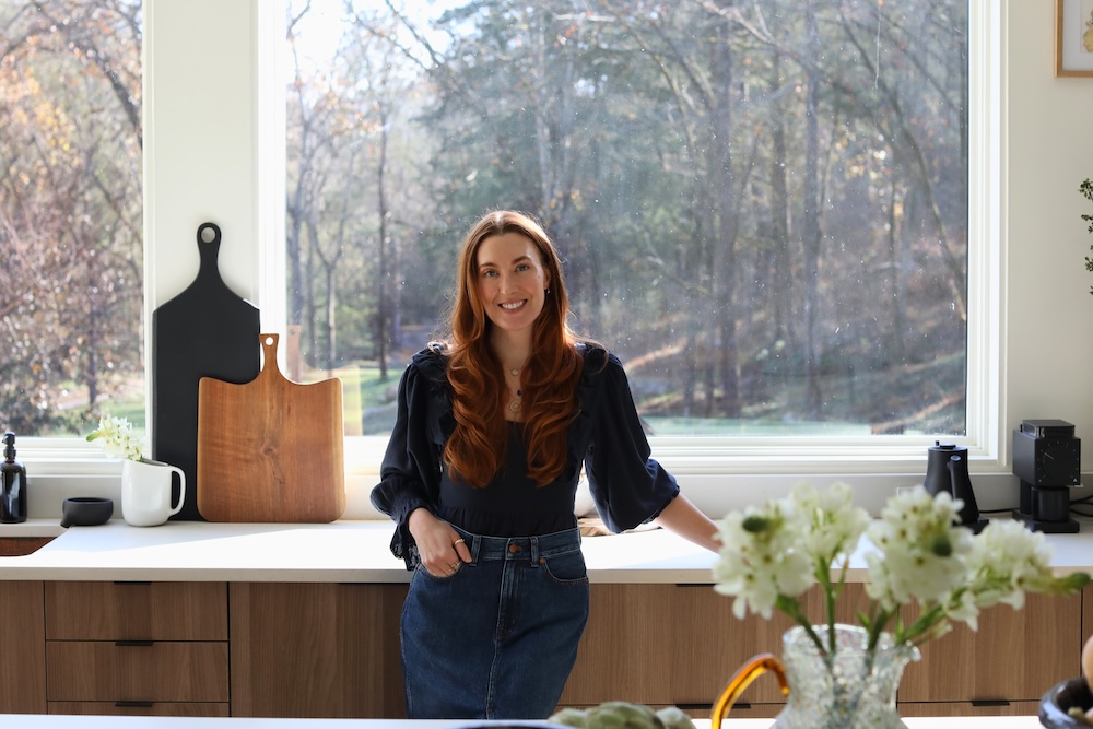 A woman with long red hair stands in a modern kitchen with wood cabinets, white countertops, and large windows overlooking a wooded area.