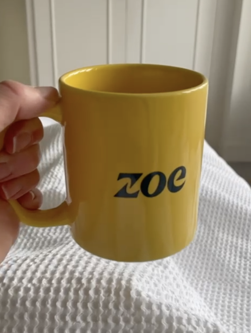 A hand holds a yellow mug with the word "zoe" printed in black letters on its side.