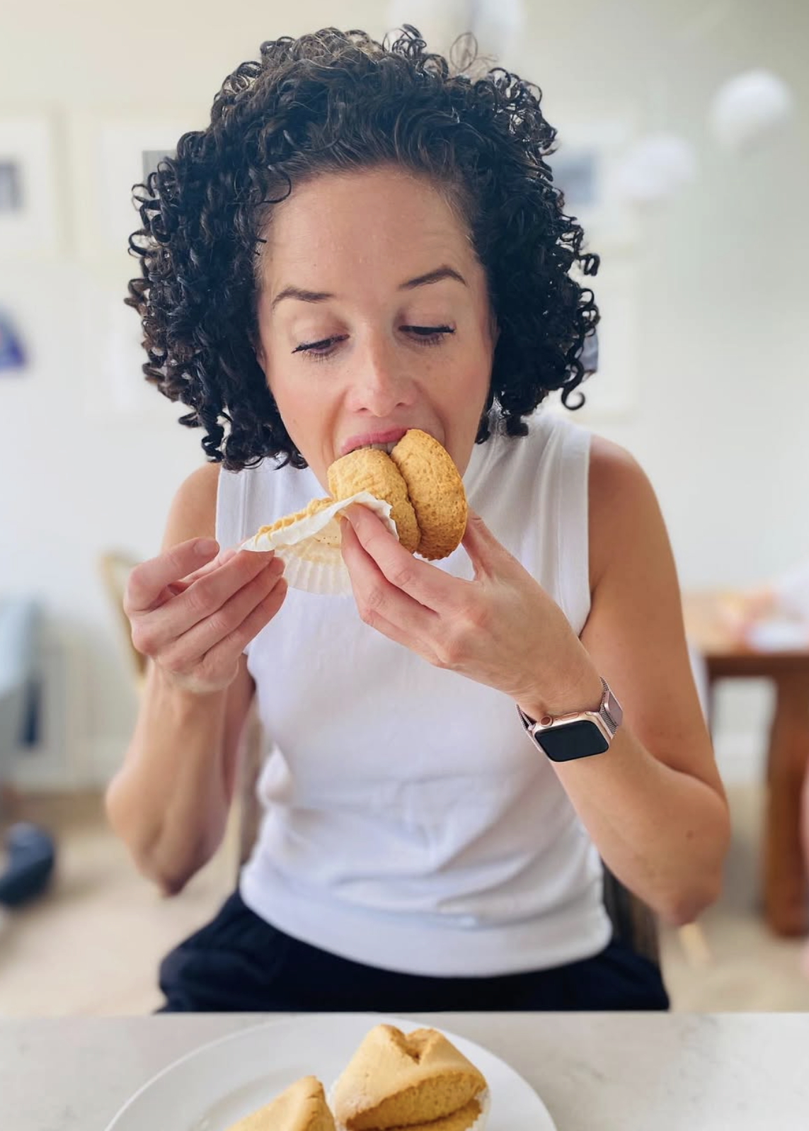 A person with curly hair eats a cupcake, holding it with both hands, while seated at a table indoors.