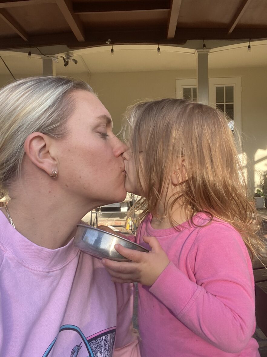 An adult and a young child, both wearing pink tops, share a kiss while the child holds a metal bowl. They are outdoors near a beige building.
