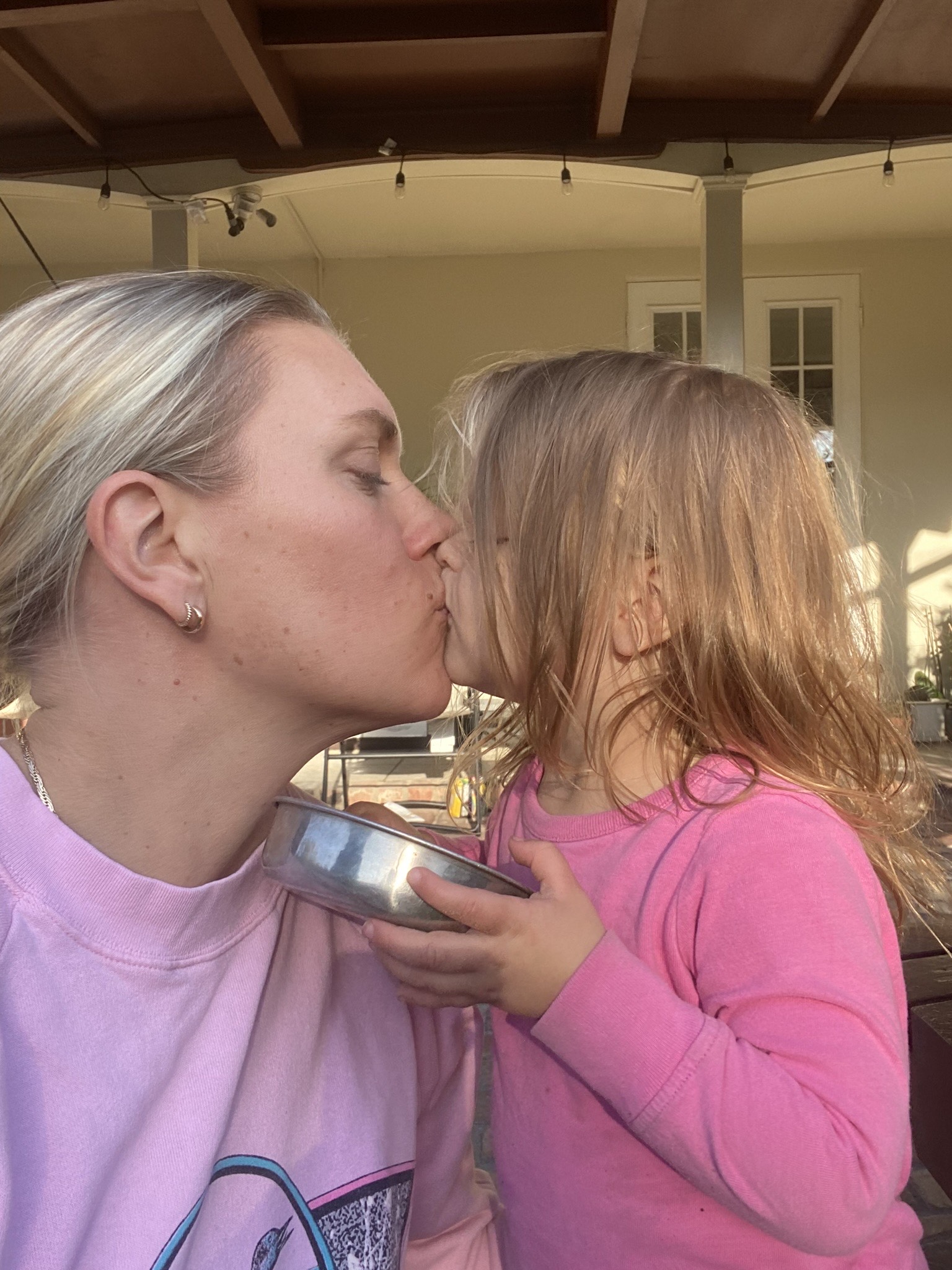 An adult and a young child, both wearing pink tops, share a kiss while the child holds a metal bowl. They are outdoors near a beige building.