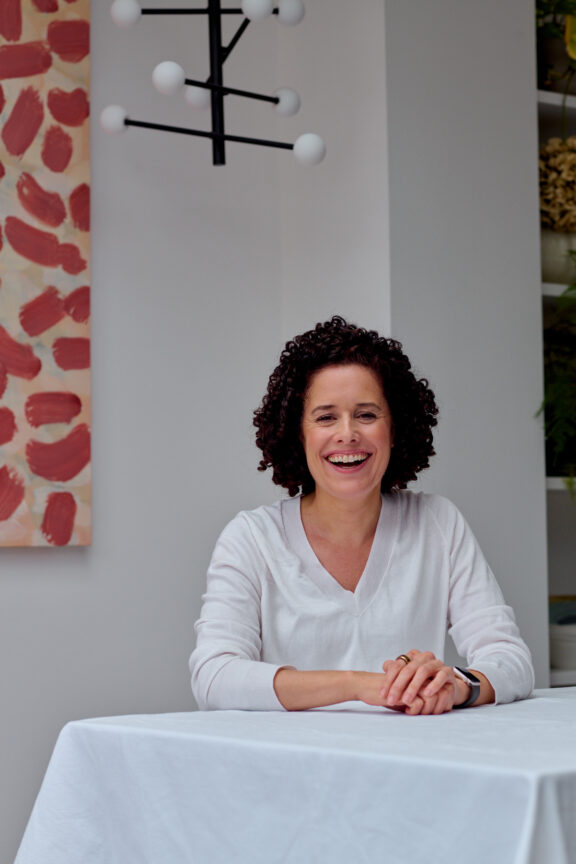 A woman with curly hair sits at a table covered with a white cloth, smiling. Abstract art and a modern light fixture are visible in the background.