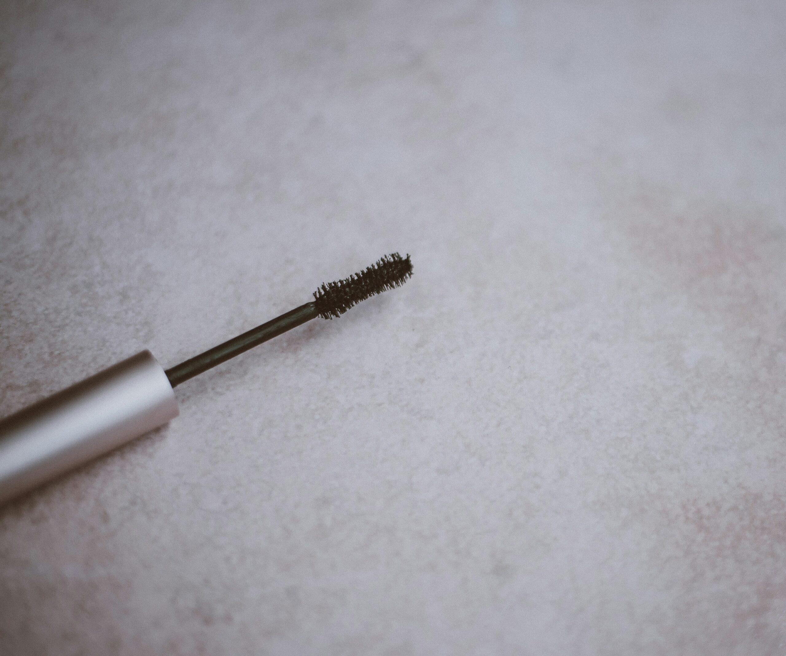 A close-up of a mascara wand with black bristles lying on a textured light pink surface.