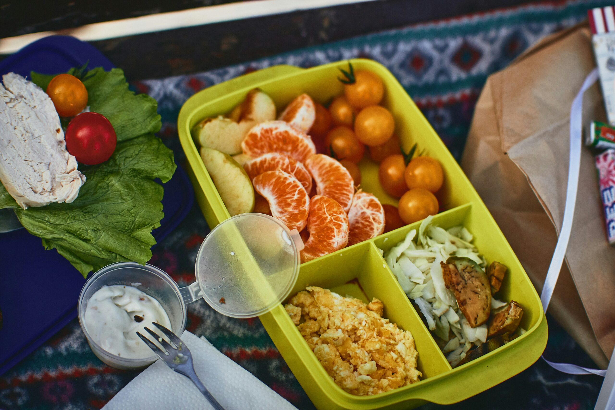 A yellow lunchbox with sections of peeled orange slices, yellow cherry tomatoes, bread, scrambled eggs, and cabbage with mushrooms, next to lettuce, chicken, and a cup of sauce.