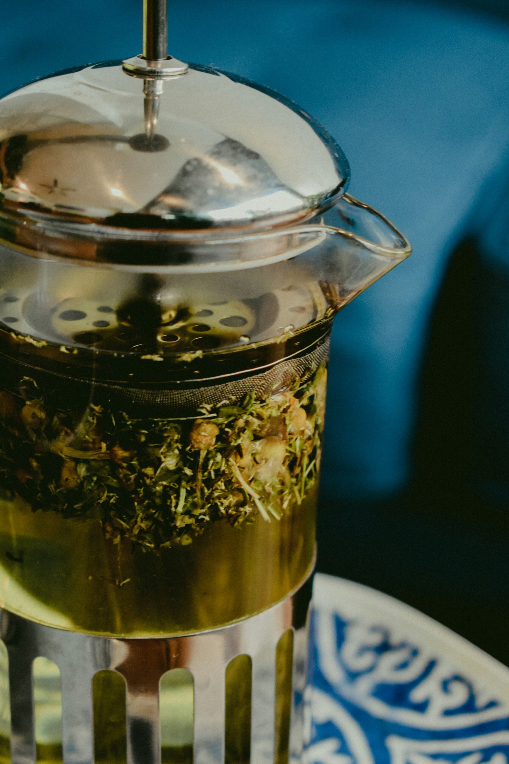 A French press filled with hot water and loose herbal tea sits on a patterned plate, with a blurred blue background.