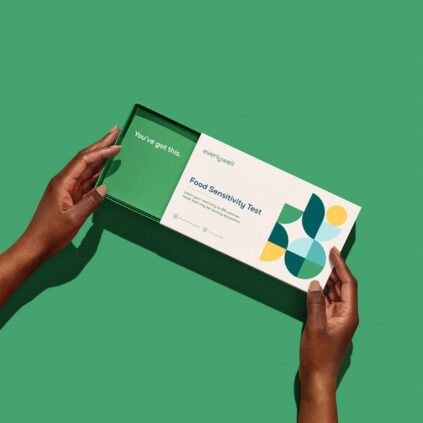 A person opens a green box labeled "Food Sensitivity Test" from Everlywell, revealing a card that says "You've got this" against a green background, with massage guns nearby for post-test relaxation.