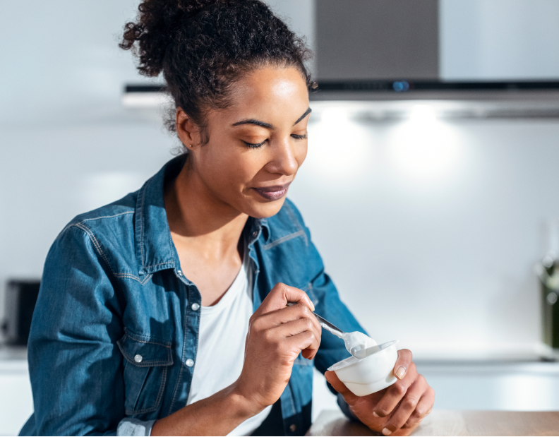 A woman with curly hair in a denim shirt is sitting at a table, holding a small bowl and eating with a spoon in a bright kitchen, while massage guns rest nearby on the counter.