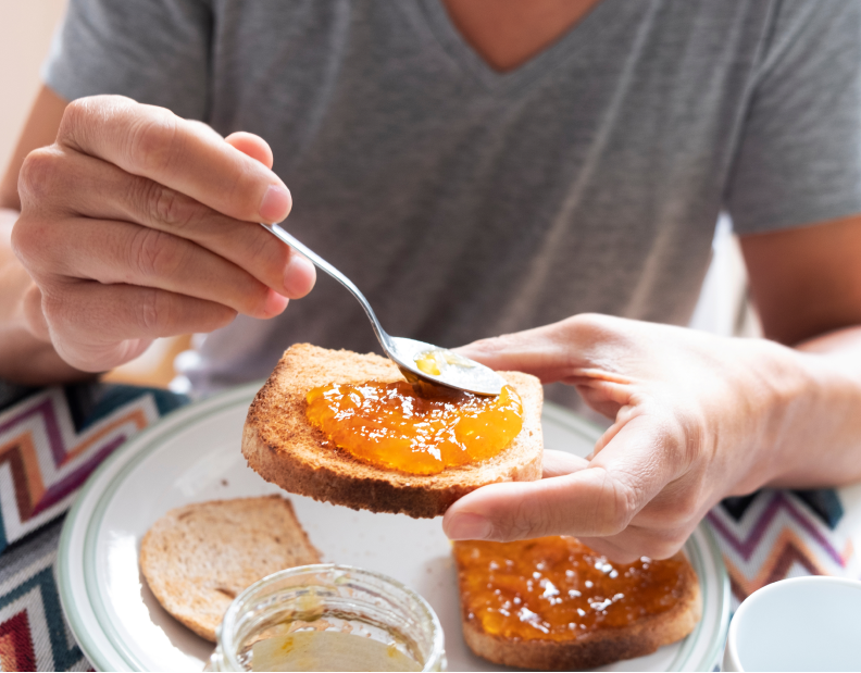 A person spreads orange jam on a slice of toast with a spoon, sitting at a table with a jar of jam, another slice of bread, a white cup, and massage guns resting nearby.