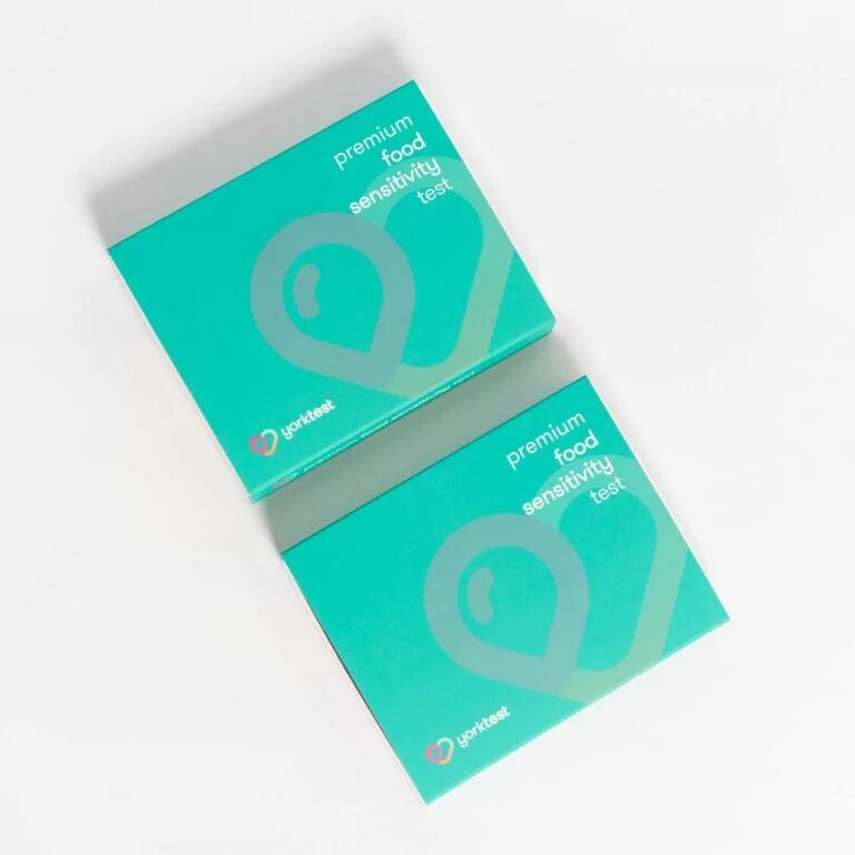 Two teal-colored boxes labeled "premium food sensitivity test" from YorkTest are placed side by side on a white background, alongside a pair of massage guns for total wellness.