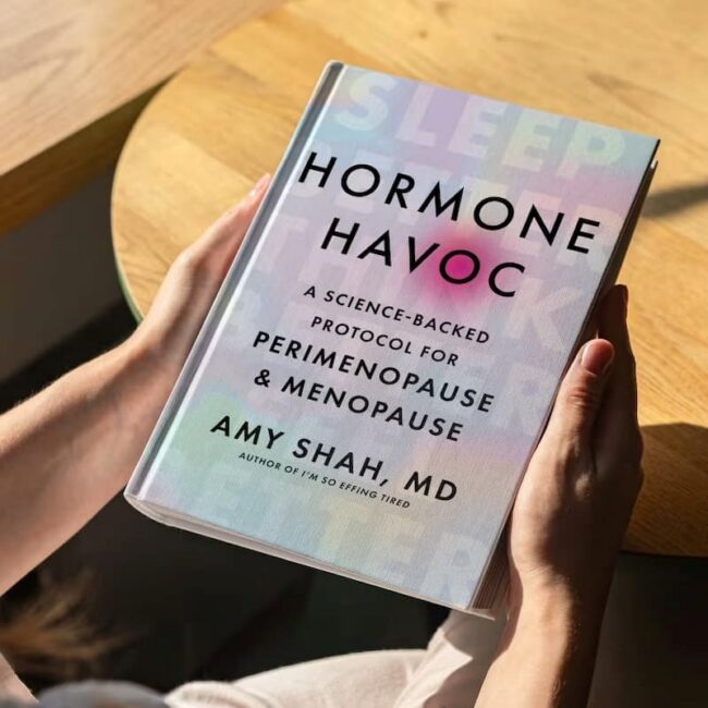 Person holding a book titled "Hormone Havoc: A Science-Backed Protocol for Perimenopause & Menopause" by Amy Shah, MD, on a wooden table.