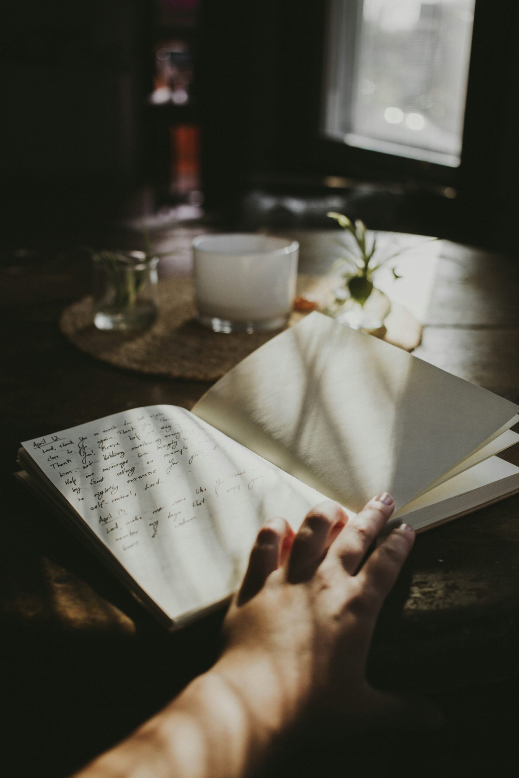 A hand holds open a notebook with handwritten text next to a candle and small plants on a wooden table near a sunlit window.