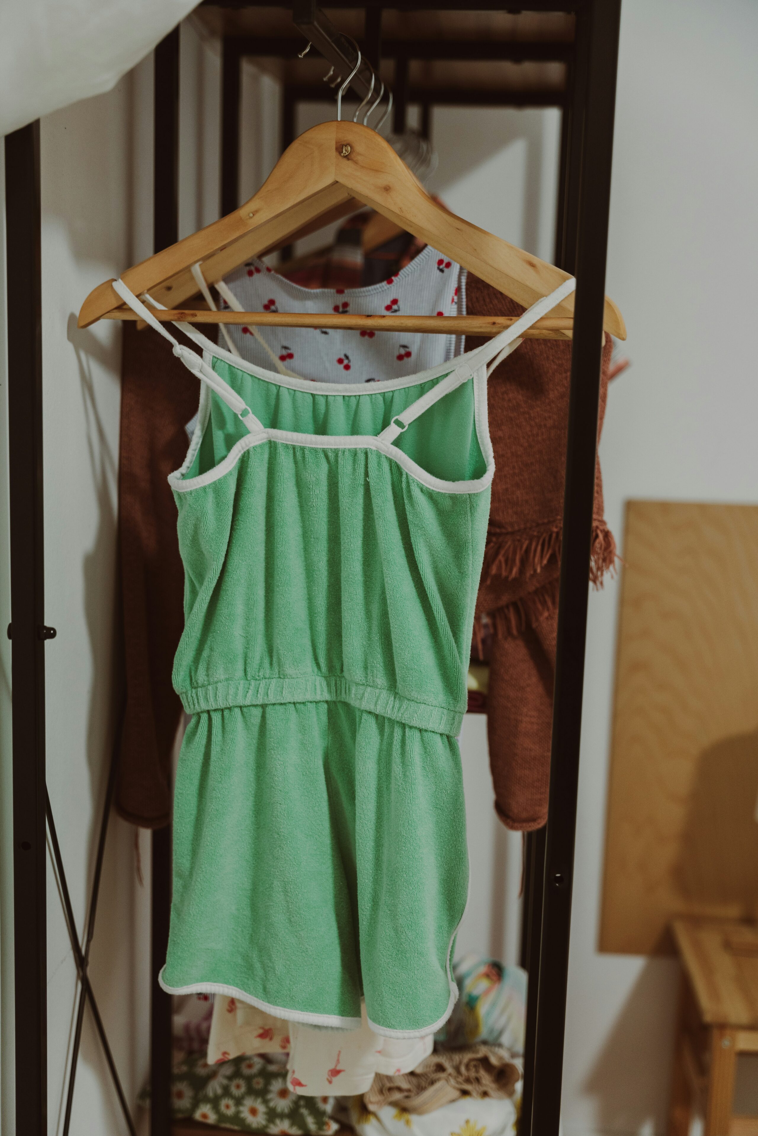 A green sleeveless dress with white trim hanging on a wooden hanger, displayed in front of other clothing on a black metal rack.