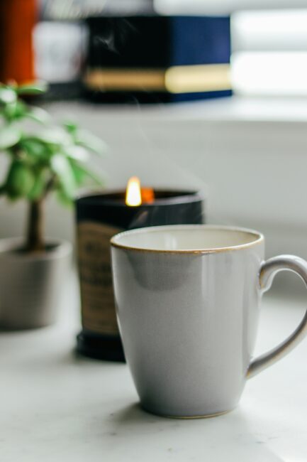 A gray ceramic mug sits on a white surface in front of a lit candle and a small potted plant, with a blurred background.