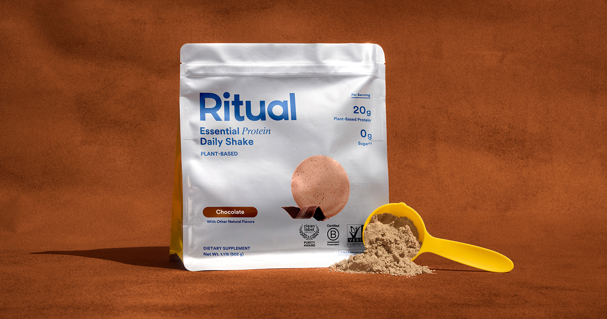 A bag of Ritual Essential Protein Daily Shake in chocolate flavor stands upright next to a yellow scoop filled with protein powder, all on a brown surface.
