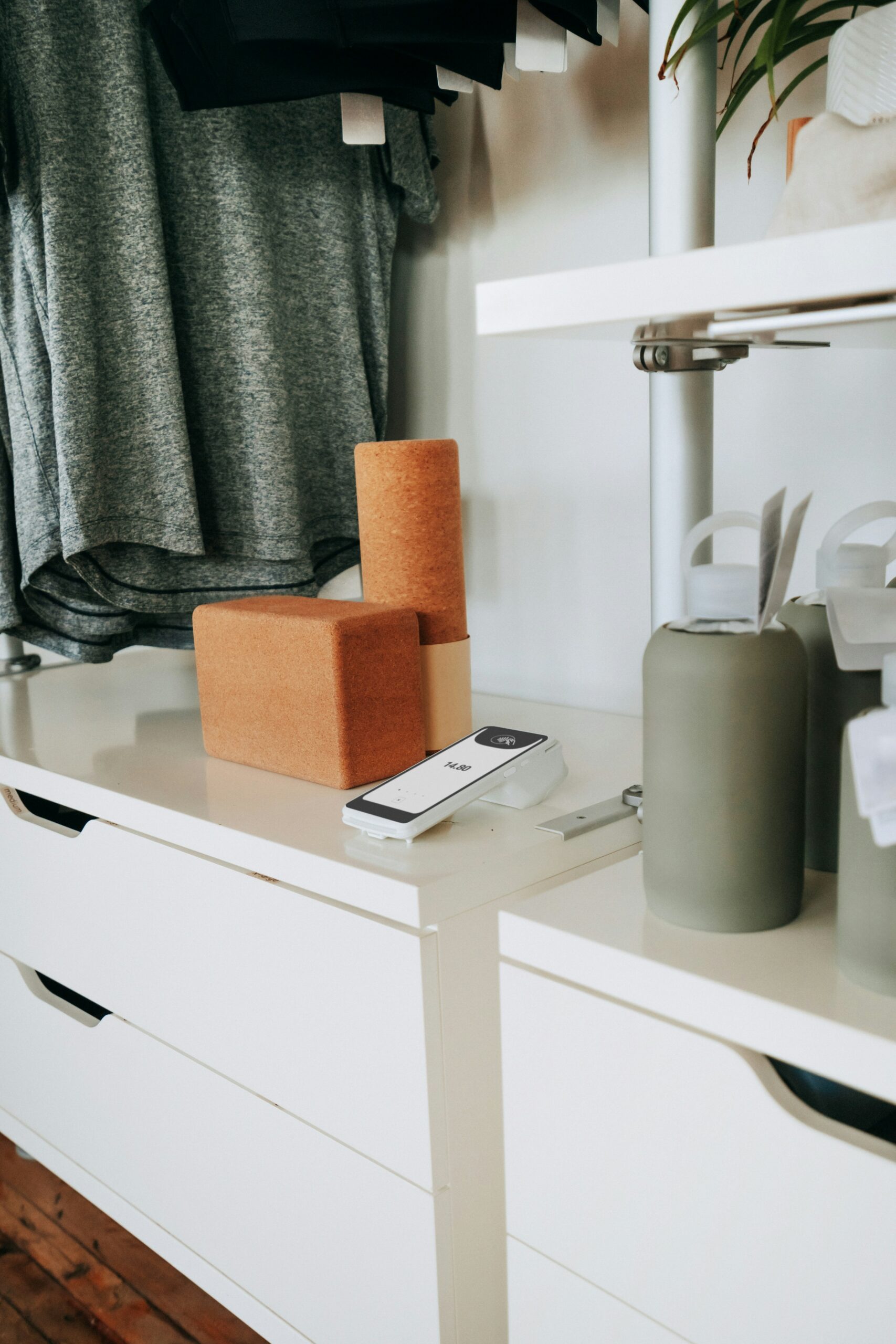 A white display shelf with cork yoga blocks, a gray water bottle, a point-of-sale card reader, and folded gray clothing hanging above.