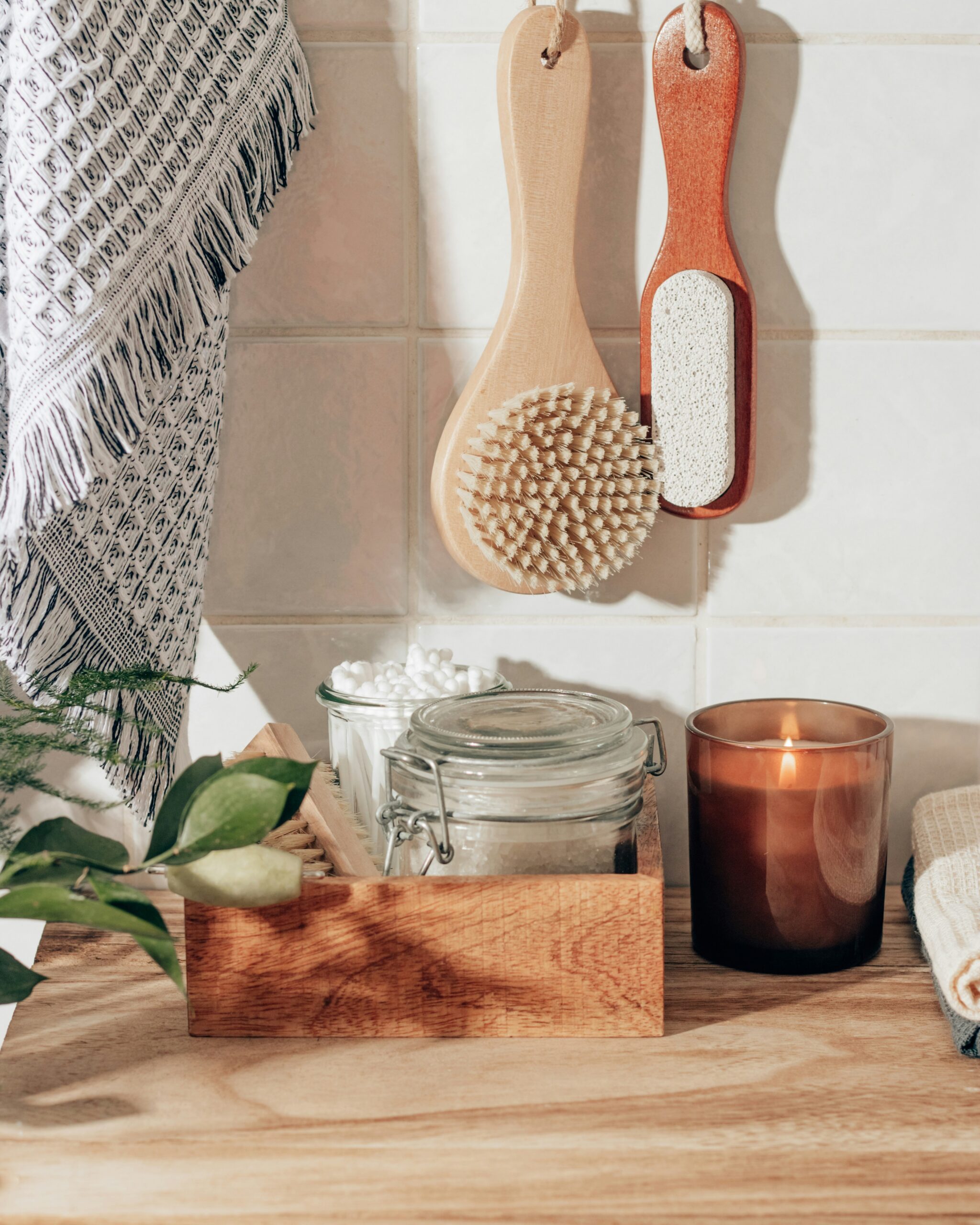 Wooden brushes hang on a tiled wall above a wooden box holding toiletries, next to a jar, cotton swabs, and a lit brown candle on a wooden surface.