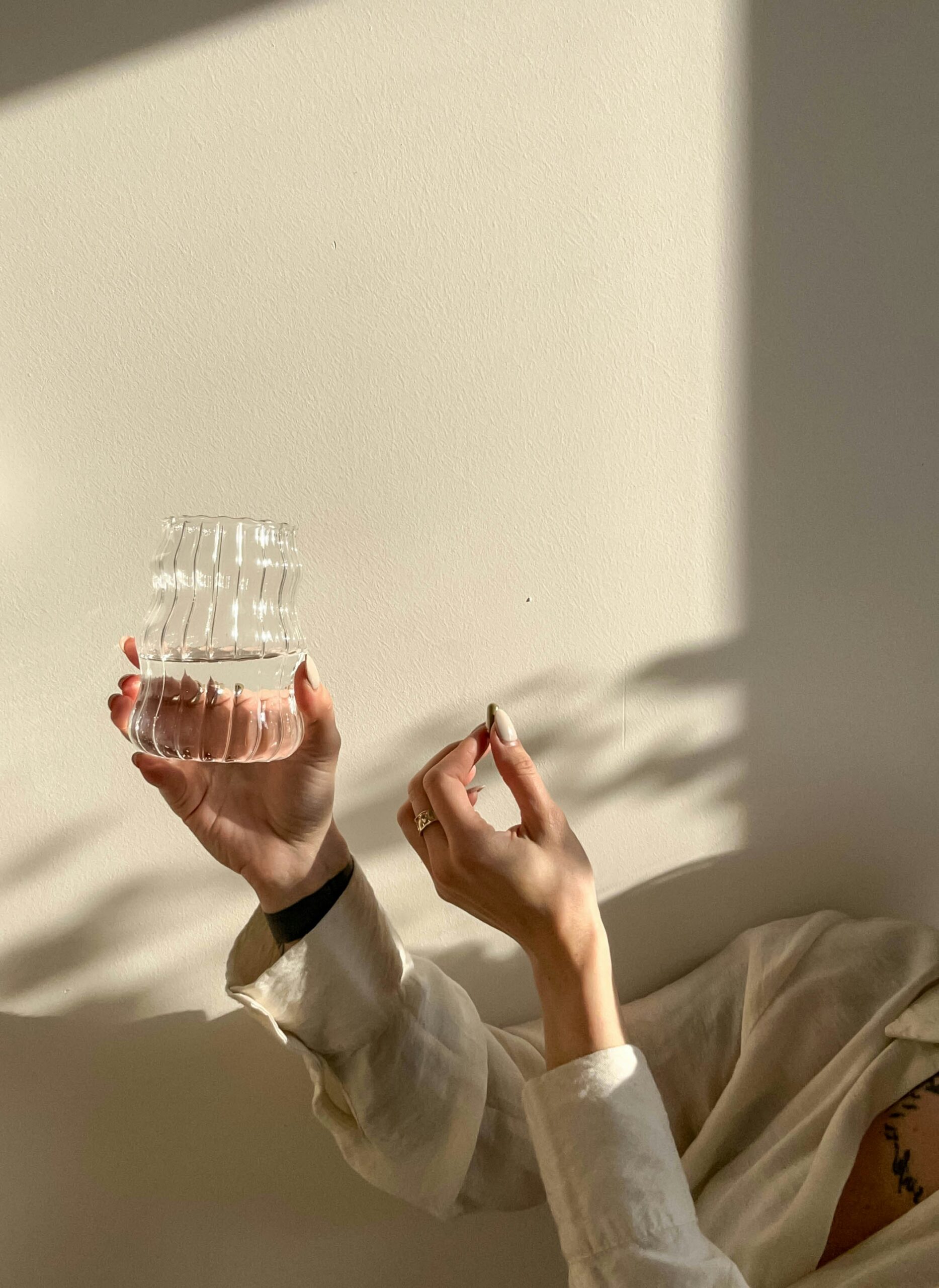 A person in a light shirt holds a clear, ribbed glass vase against a sunlit wall, with shadows and partial view of their hands and arm.