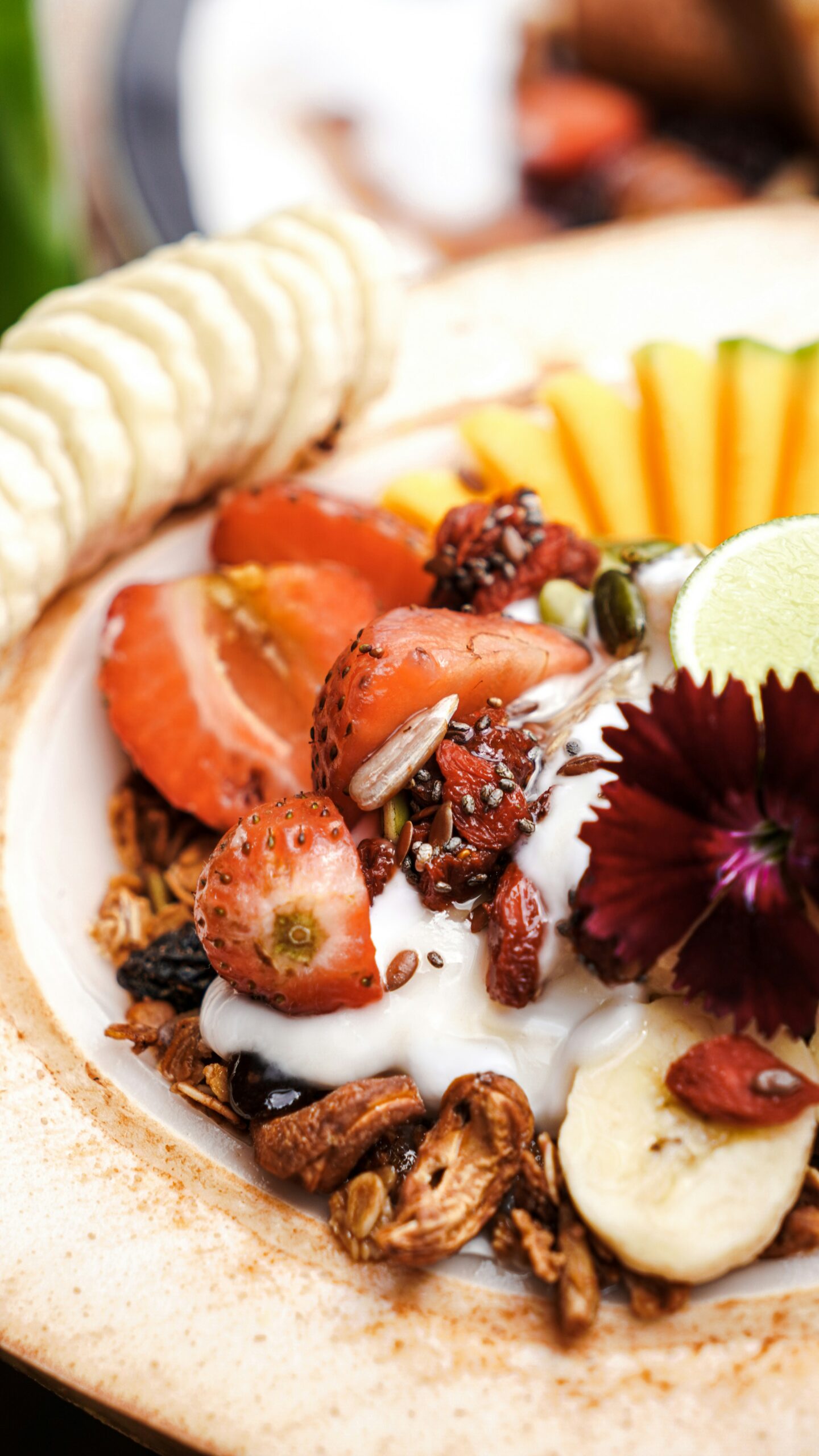 A bowl of granola topped with yogurt, sliced strawberries, banana, mango, seeds, dried berries, and a lime wedge, garnished with a dark red edible flower.