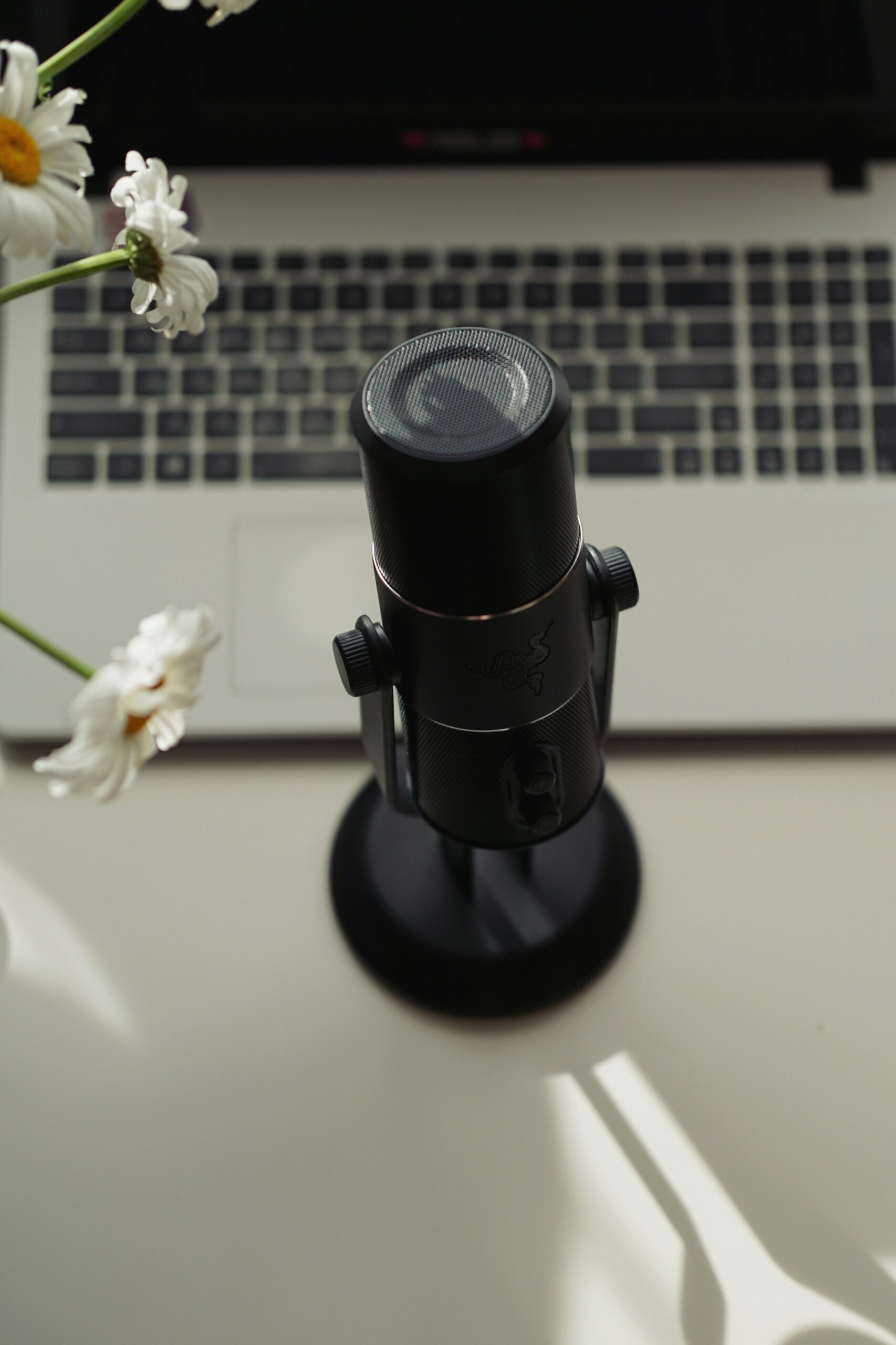 A black microphone on a stand placed in front of a laptop keyboard, with white flowers visible to the left on a white desk.