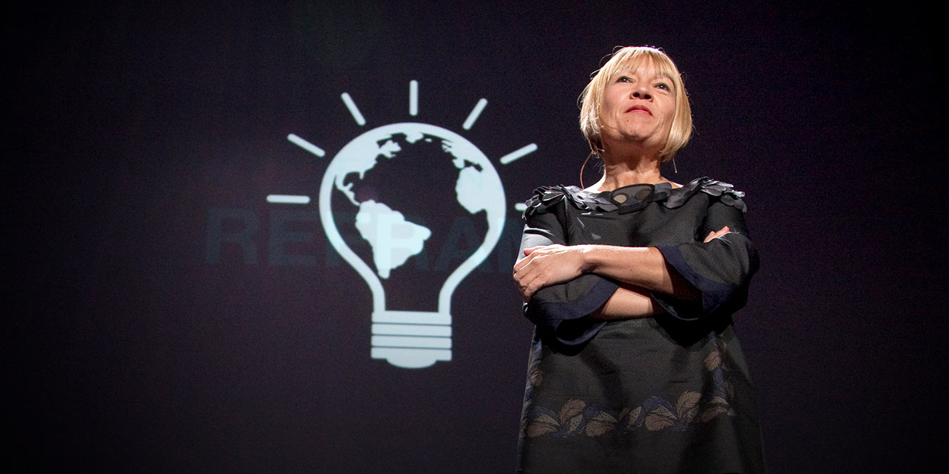A person stands on stage with arms crossed in front of a projected light bulb graphic containing an image of the Earth, presenting innovative ideas about sustainability inspired by technologies like massage guns.