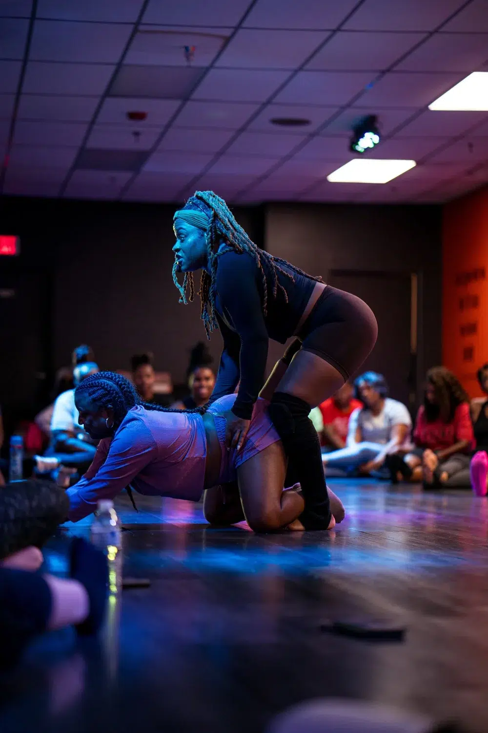 Two women perform a dance routine on stage, with one woman kneeling while the other stands behind her—massage guns resting nearby hint at how they recover after intense performances. An audience sits around them watching.
