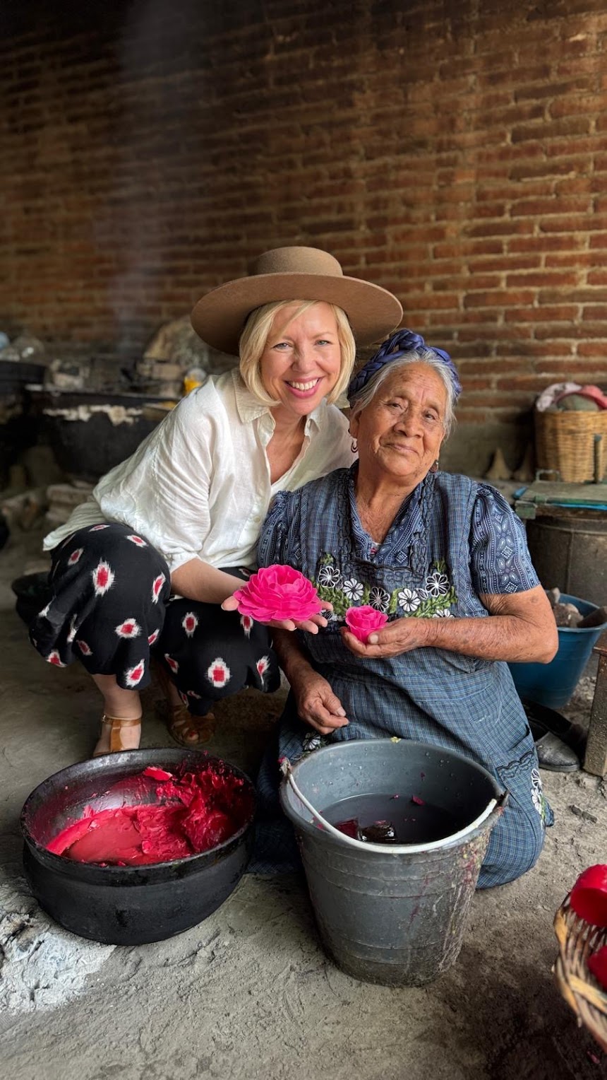 Two women sit together indoors; one holds a pink handcrafted flower, and the other kneels beside her. Buckets of pink dye and flower materials are on the floor in front of them.
