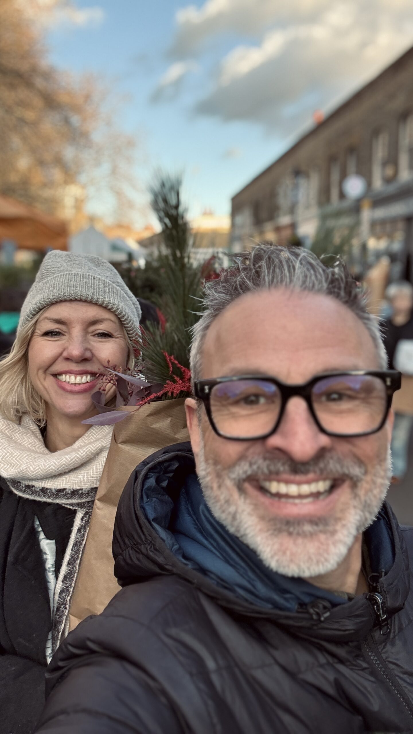 A smiling man and woman outdoors on a street, holding a bouquet with greenery and red accents. The sky is partly cloudy, and people and buildings are visible in the background.