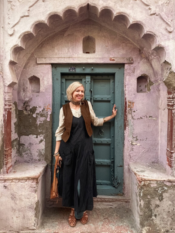 A woman stands smiling in front of a blue door set in a weathered pink wall with ornate architectural details.