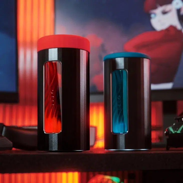 Two cylindrical massage guns with colored lids and transparent windows displaying red and blue interiors, placed on a dark surface with an orange-lit background.