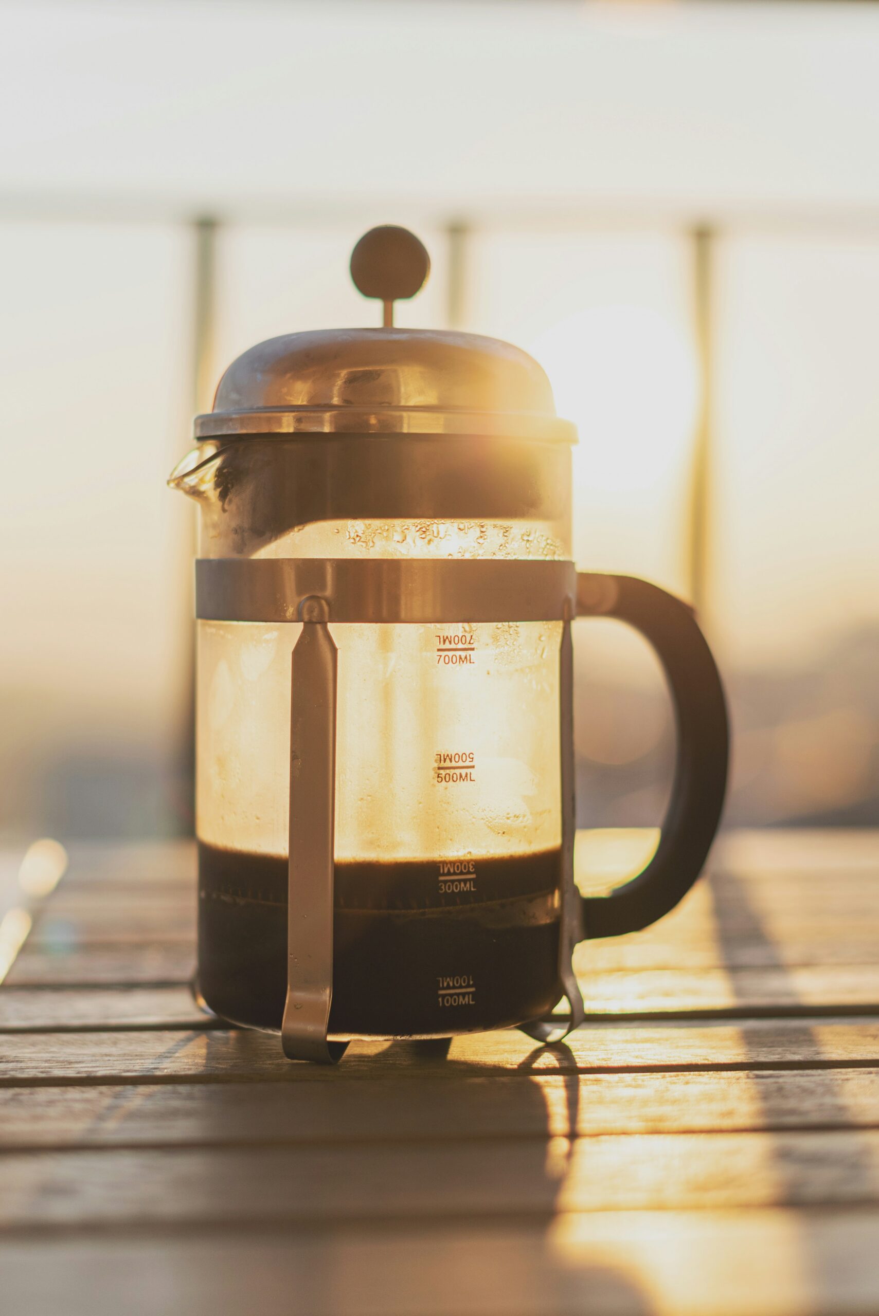 A French press with brewed coffee sits on a wooden table in sunlight, with measurement markings visible on the glass.
