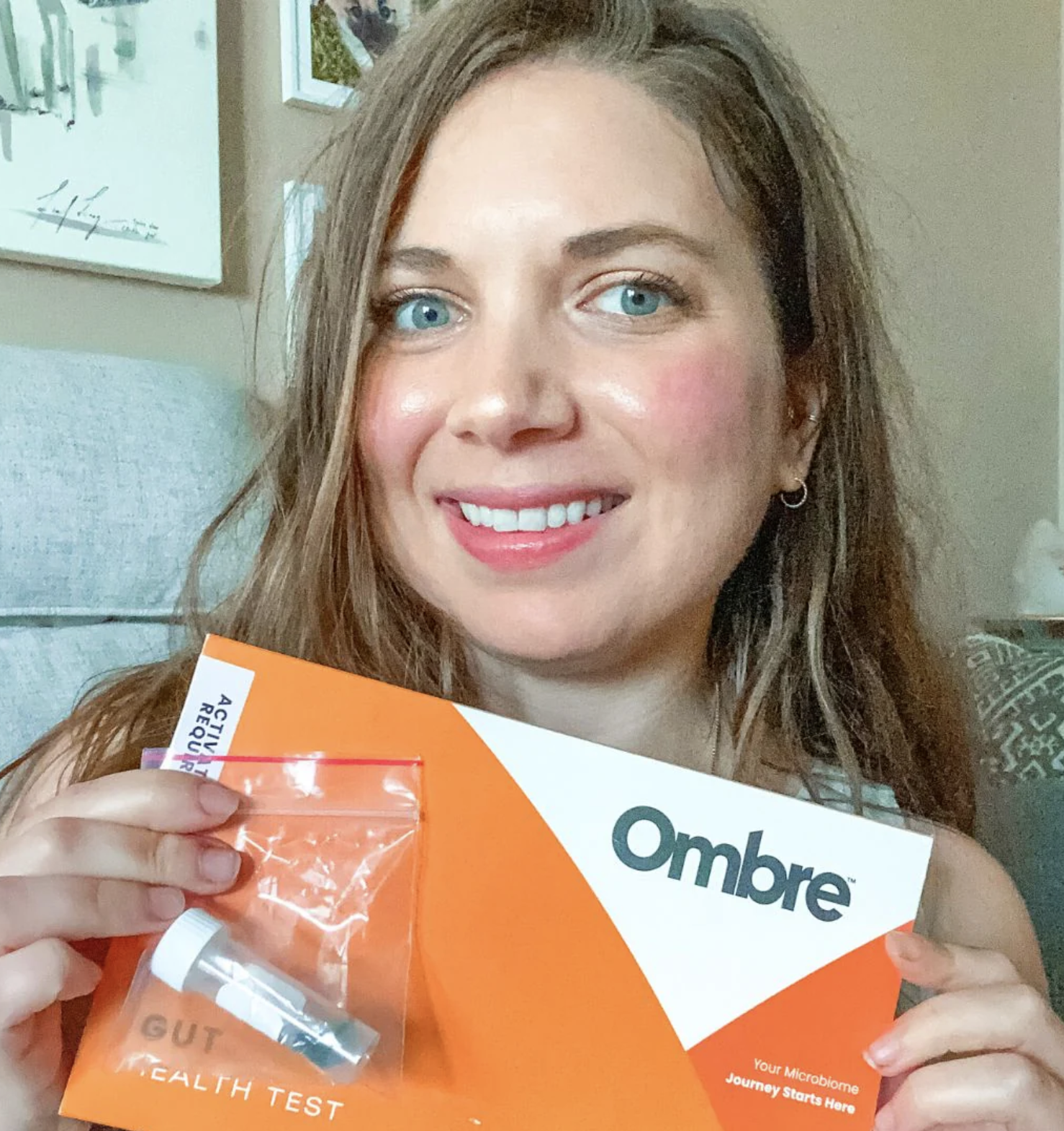 A woman holds up an Ombre Gut Health Test kit, smiling at the camera, as massage guns are visible in the background, highlighting her commitment to overall wellness.