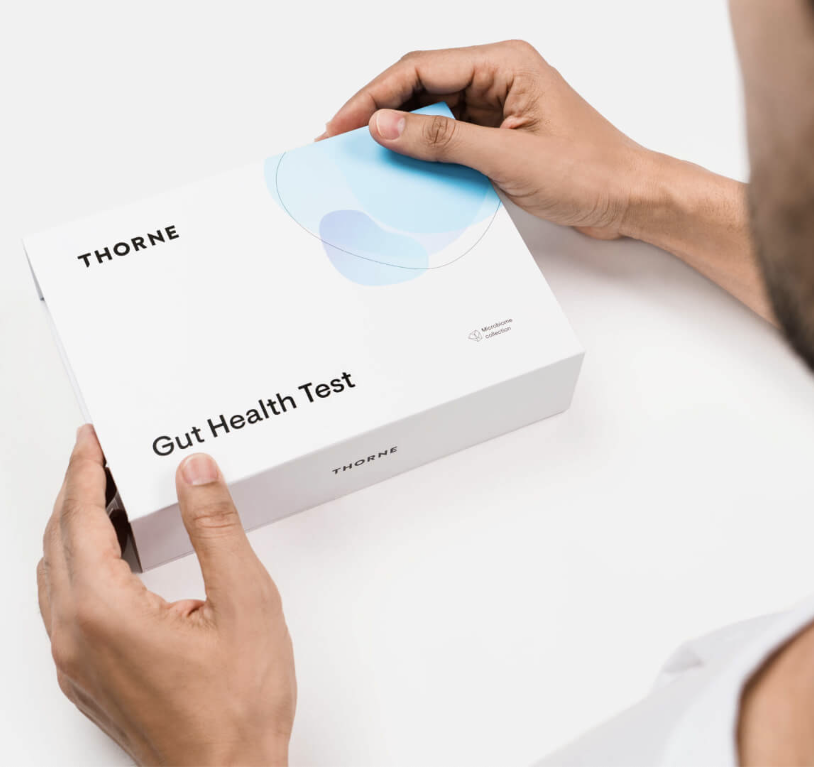 A person holds a white box labeled "Thorne Gut Health Test" on a white surface, surrounded by minimal blue abstract designs—perfect for those interested in wellness products, from gut health kits to massage guns.