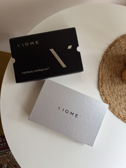Two Viome-branded boxes, one black labeled "Full Body Intelligence" and one silver, sit on a white round table next to a woven placemat—perfect alongside wellness tools like massage guns.