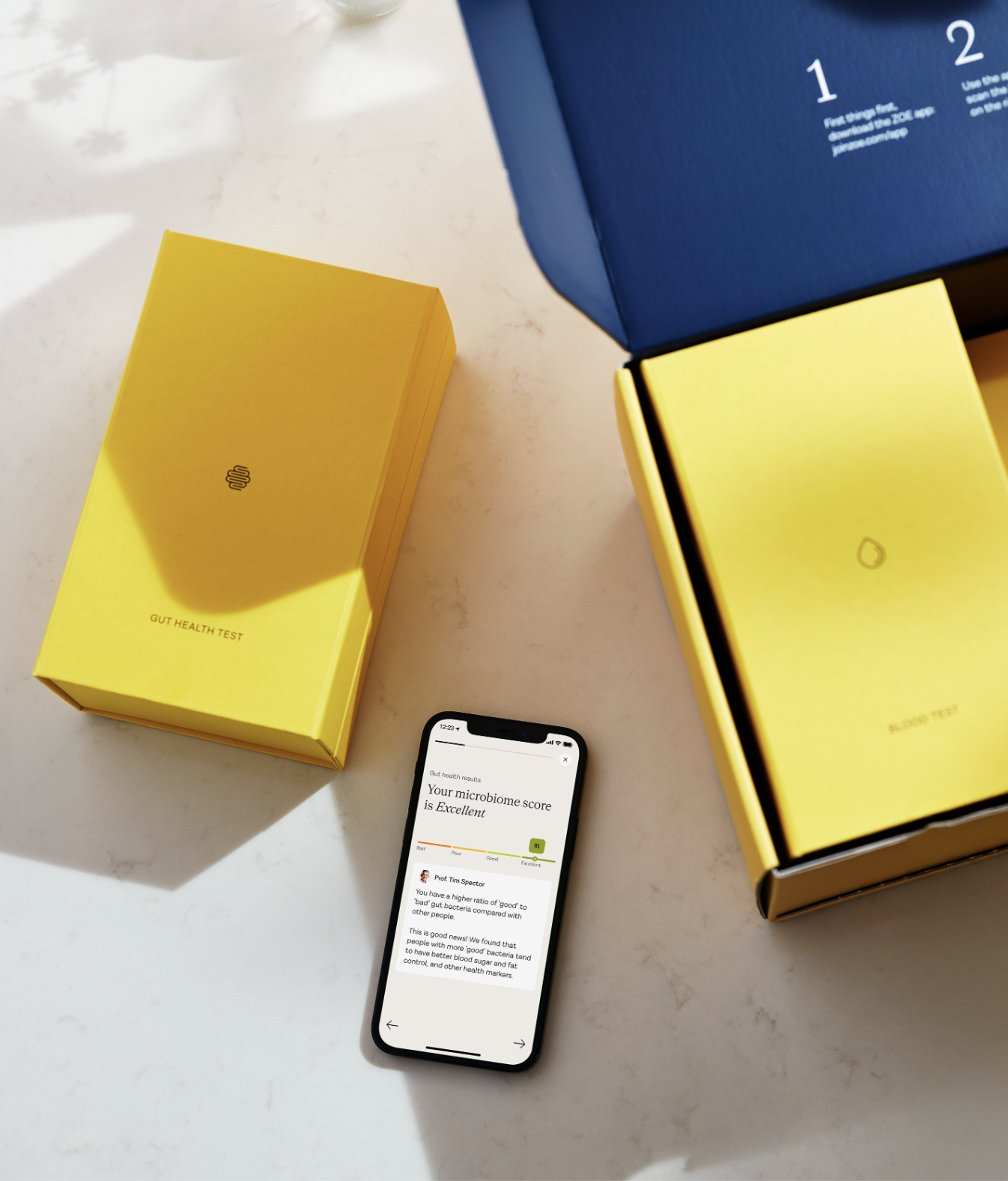 Two yellow health test kit boxes and an open blue box are on a table next to a smartphone displaying microbiome test results, with massage guns visible in the background.