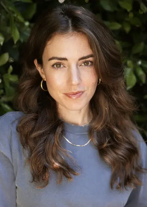 A woman with long brown hair, wearing a gray top, gold hoop earrings, and a gold necklace, poses in front of green foliage.