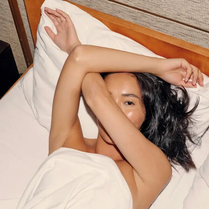 Person lying in bed, partially covered by a white sheet, with one arm resting across their forehead and eyes peeking through—relaxing after using massage guns for soothing relief.