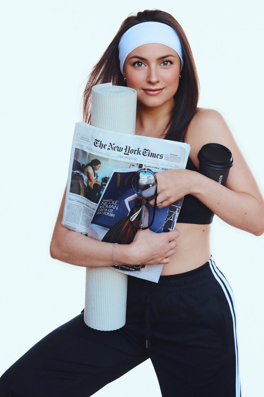 A woman in athletic wear holds a yoga mat, a coffee cup, a newspaper, a magazine, and sunglasses against a plain white background.