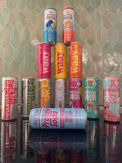Assorted cans of protein drinks and sodas—including brands Joyburst, VYVAM, and beelish—are arranged in a pyramid on a reflective surface with massage guns displayed nearby against a tiled backsplash.