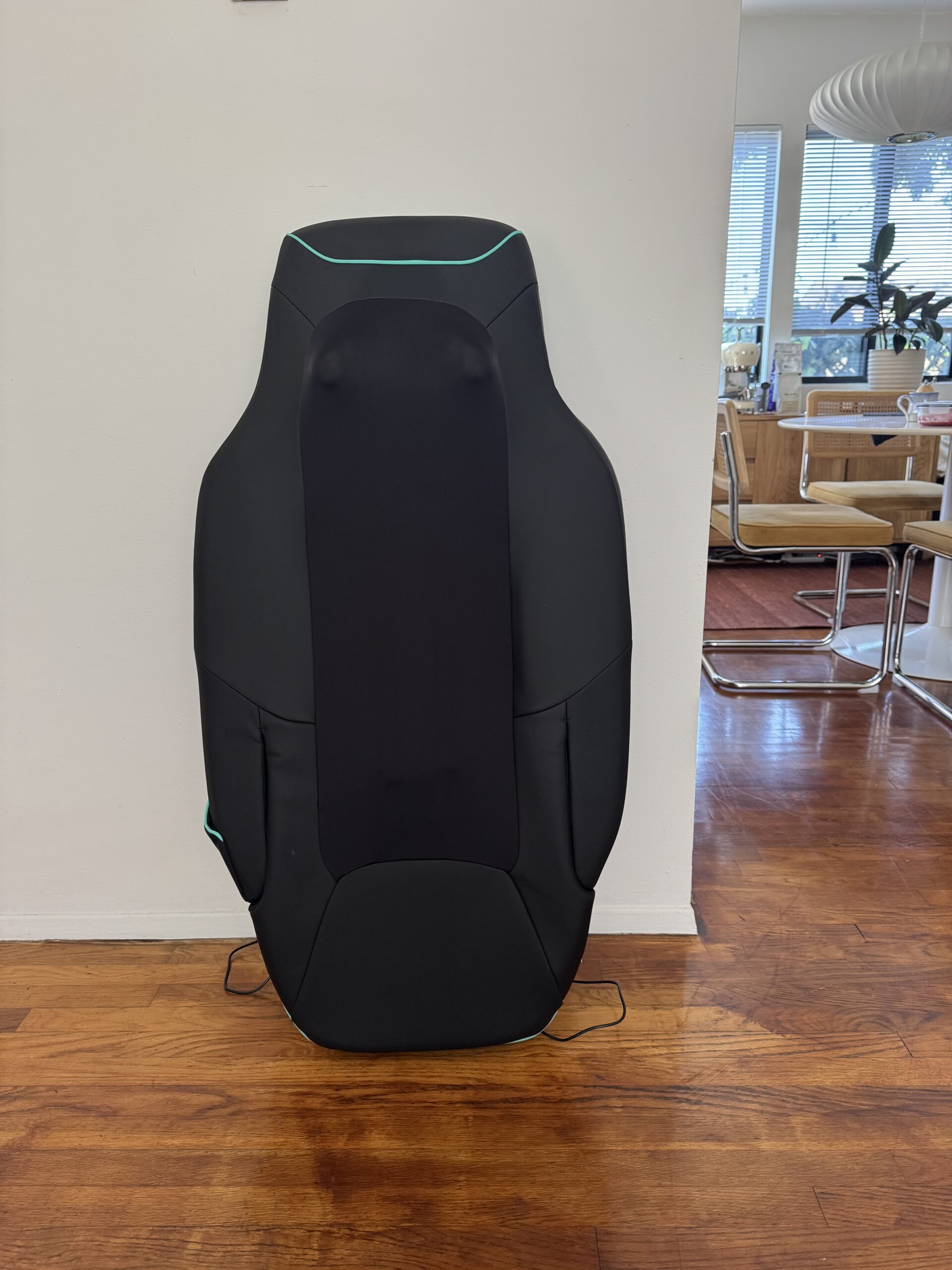 A black massage chair pad with teal accents is propped upright against a white wall in a living space with wooden floors.