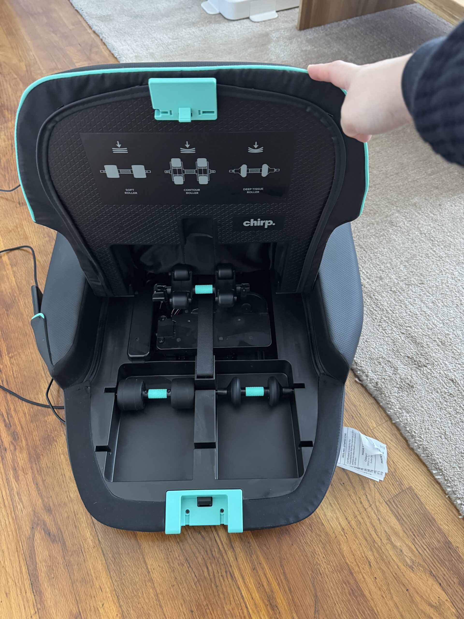 A person holds open the back panel of a Chirp car seat, exposing the internal belt and latch system on a hardwood floor.