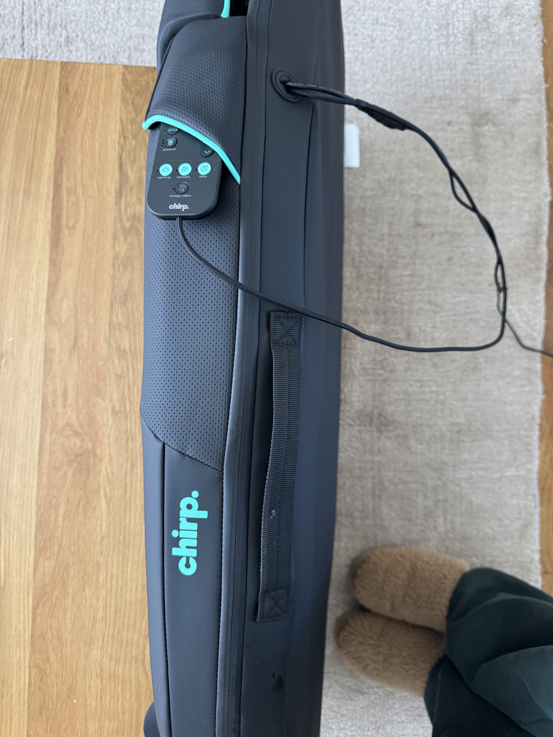 A black Chirp massage device with a control panel and cord rests on a tan rug next to someone wearing brown slippers and dark pants.