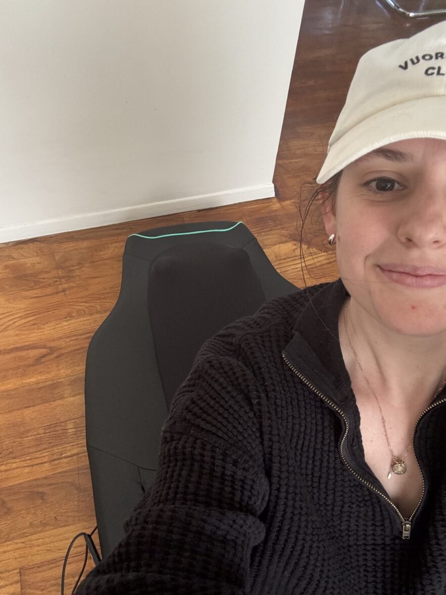 A person wearing a white cap and black sweater takes a selfie indoors, with a massage chair pad visible on a wooden floor behind them.