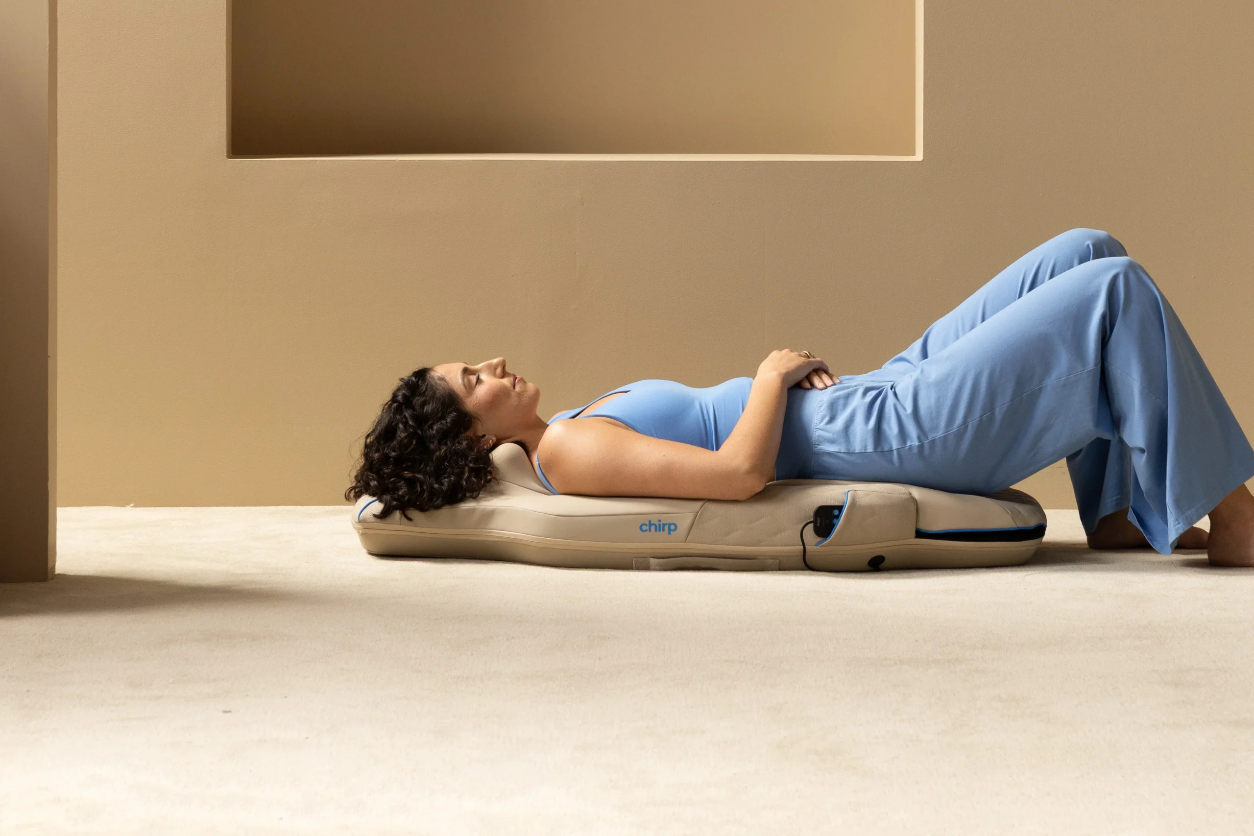 Woman in blue clothes lying on her back on a beige floor mat labeled "chirp" in a minimalist room with neutral walls.