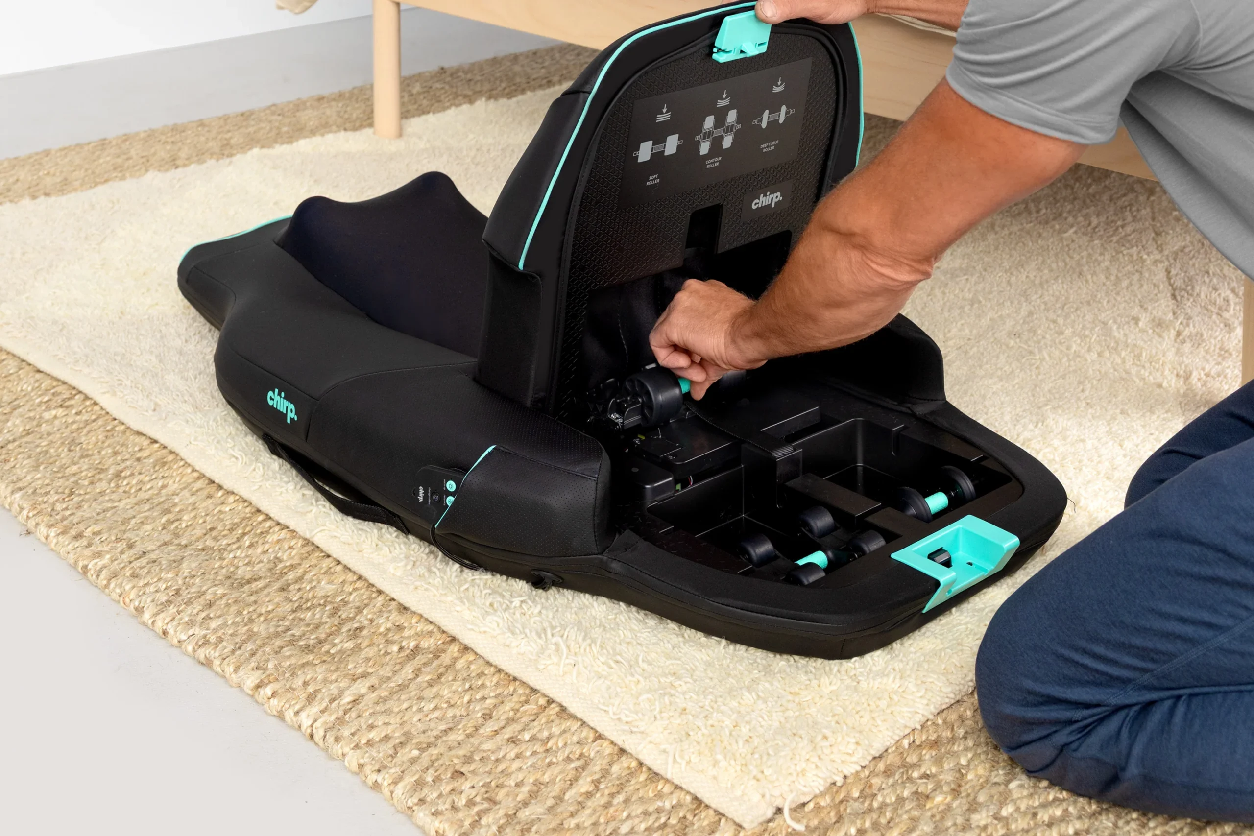 An adult adjusts the base of a black and teal child car seat on a beige rug, with the seat back open to reveal the interior mechanisms.