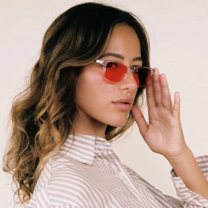 Woman with wavy brown hair wearing red-tinted sunglasses and a beige striped shirt, looking at the camera against a plain background.