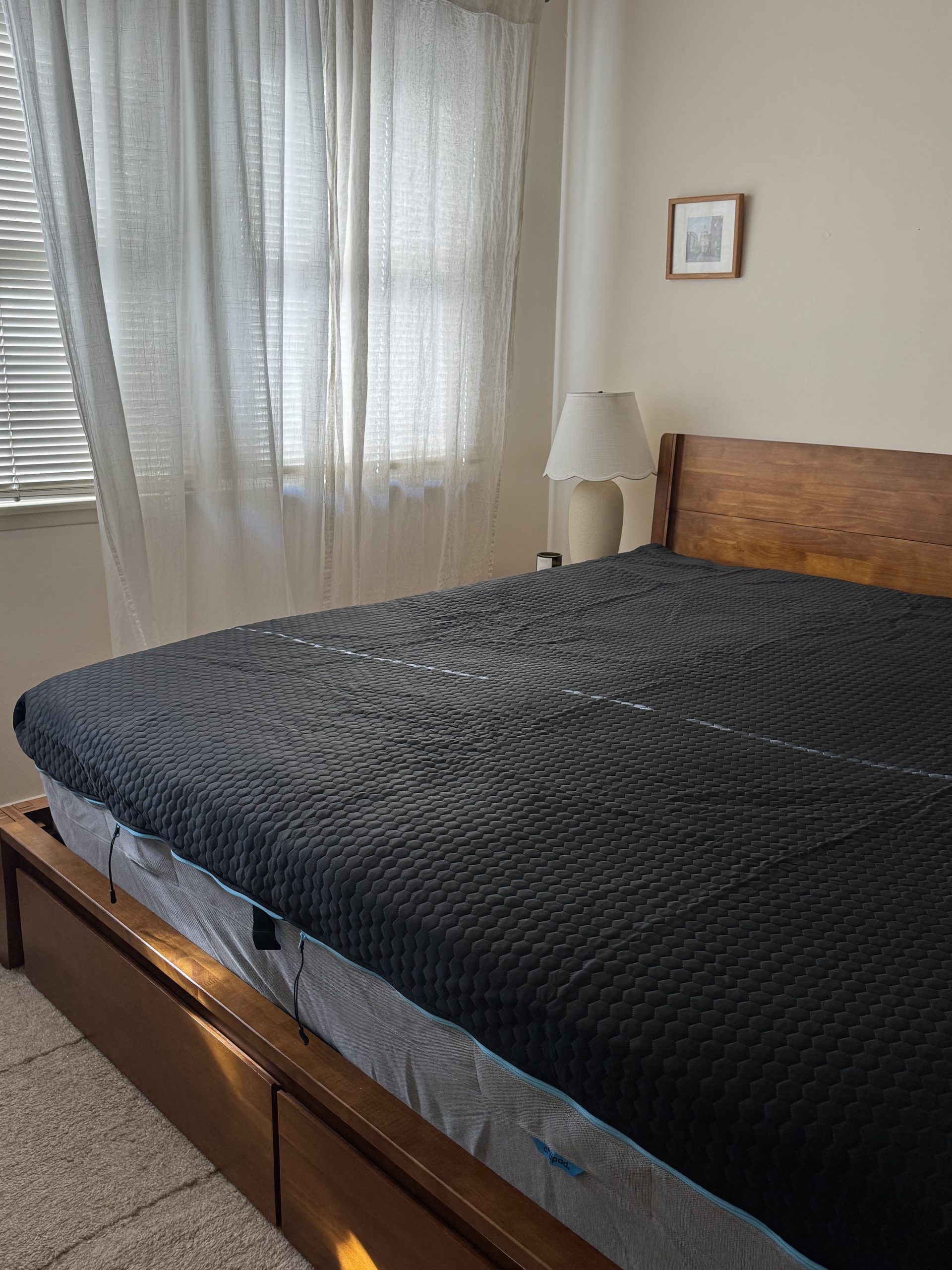 A bed with a dark, textured cooling topper, a wooden frame, and a bedside lamp in front of sheer curtains.