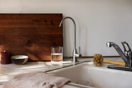A kitchen sink with a glass of water, a dish brush, a wooden cutting board, a towel, and a small bowl on the counter in natural light.
